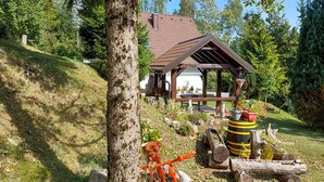 Un chalet rustique doté d’une terrasse en bois et d’un espace extérieur couvert, entouré d’une végétation luxuriante et bordé par une forêt.