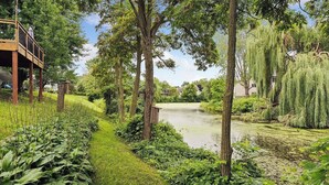 Un jardí tranquil al costat del riu, amb terrassa de fusta, vegetació abundant i arbres madurs.