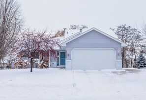 Une maison de plain-pied avec un garage, entourée d’un paysage enneigé et d’arbres sans feuilles.