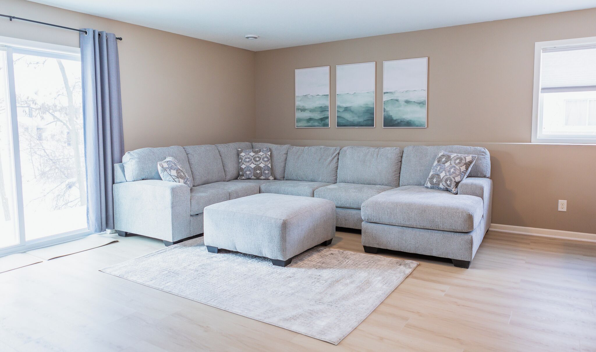 Un salon moderne avec un canapé d’angle gris, un pouf assorti et une grande fenêtre agrémentée de rideaux.