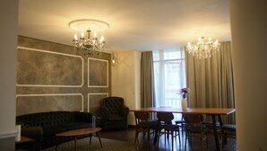 Uma sala de estar com um sofá escuro, uma mesa de centro de madeira, uma mesa de jantar com cadeiras e duas luminárias de cristal.
