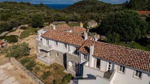 Un bâtiment à deux étages, doté d’un toit en tuiles, entouré de verdure et de collines.