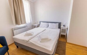Un lit double avec du linge de lit blanc et deux oreillers, une table de chevet avec une lampe, une horloge et un radiateur placé sous la fenêtre.