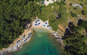 Oversigtsbillede af en strand med krystalklart turkis vand, omgivet af frodig, grøn skov. Flere rækker strandparasoller og liggestole er placeret langs kystlinjen.