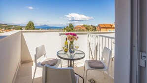 Ein Balkon mit zwei weißen Stühlen, einem kleinen runden Tisch mit Blumen und Weinflaschen, und Blick auf Meer und Berge.