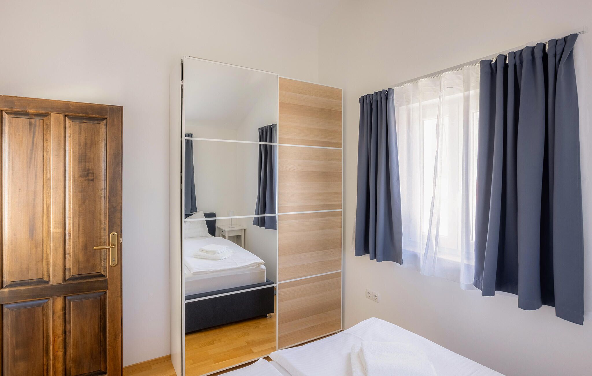 Un dormitorio con un armario de madera, una cama con ropa blanca, una mesita de noche y una ventana con cortinas oscuras.