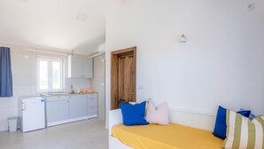 Ein kleines Zimmer mit einem Bett, einer kleinen Küchenzeile, einem Fenster und einer Tür.
