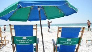 Beach chairs with "Sandpiper Cove Chair Rentals" signs, a large blue and green umbrella, and the ocean in the background. - Beachfront Bliss with Panoramic Gulf Views | Sandpiper Cove 2152 Destin, FL (Destin)