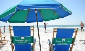 Beach chairs with "Sandpiper Cove Chair Rentals" signs, a large blue and green umbrella, and the ocean in the background. - Beachfront Bliss with Panoramic Gulf Views | Sandpiper Cove 2152 Destin, FL (Destin)