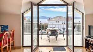 Ein Zimmer mit einer großen Glasdoor, die auf einen Balkon mit Tisch und Stühlen führt, einem Fernseher und Blick auf ein Gebäude und Wasser.