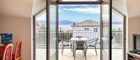 A room with a large glass door leading to a balcony with a table and chairs, a TV, and a view of a building and water.