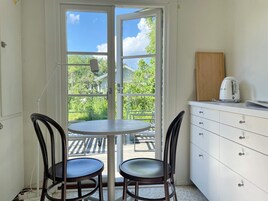 Una habitación con una mesa y dos sillas, una kitchenette con cafetera y vistas al exterior con vegetación.