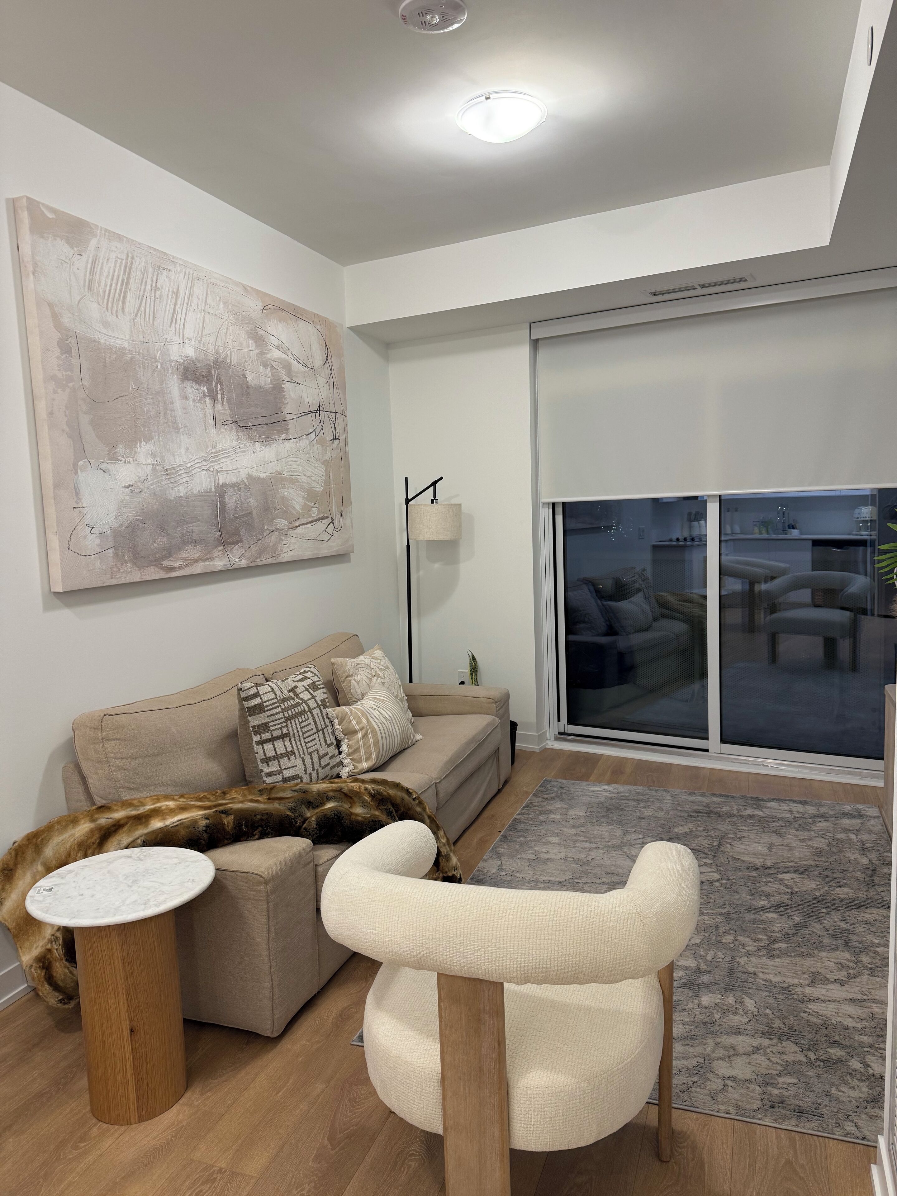 Una sala de estar moderna con un sofá color beige, una mesita redonda de madera, un sillón blanco y un cuadro abstracto grande en la pared.