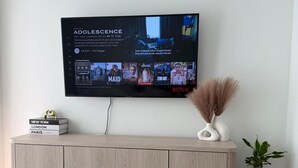 A modern living room with a flat-screen TV mounted on the wall displaying a series of shows. Below the TV, there is a light-colored, ribbed console with a few books and decorative items. - Fully Furnished Stylish Vaughan Condo near / Steps to VaughanMills (Vaughan)