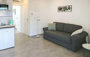 Een kleine leefruimte met een kitchenette, een grijze bank met patroon kussens, en een vloerlamp.