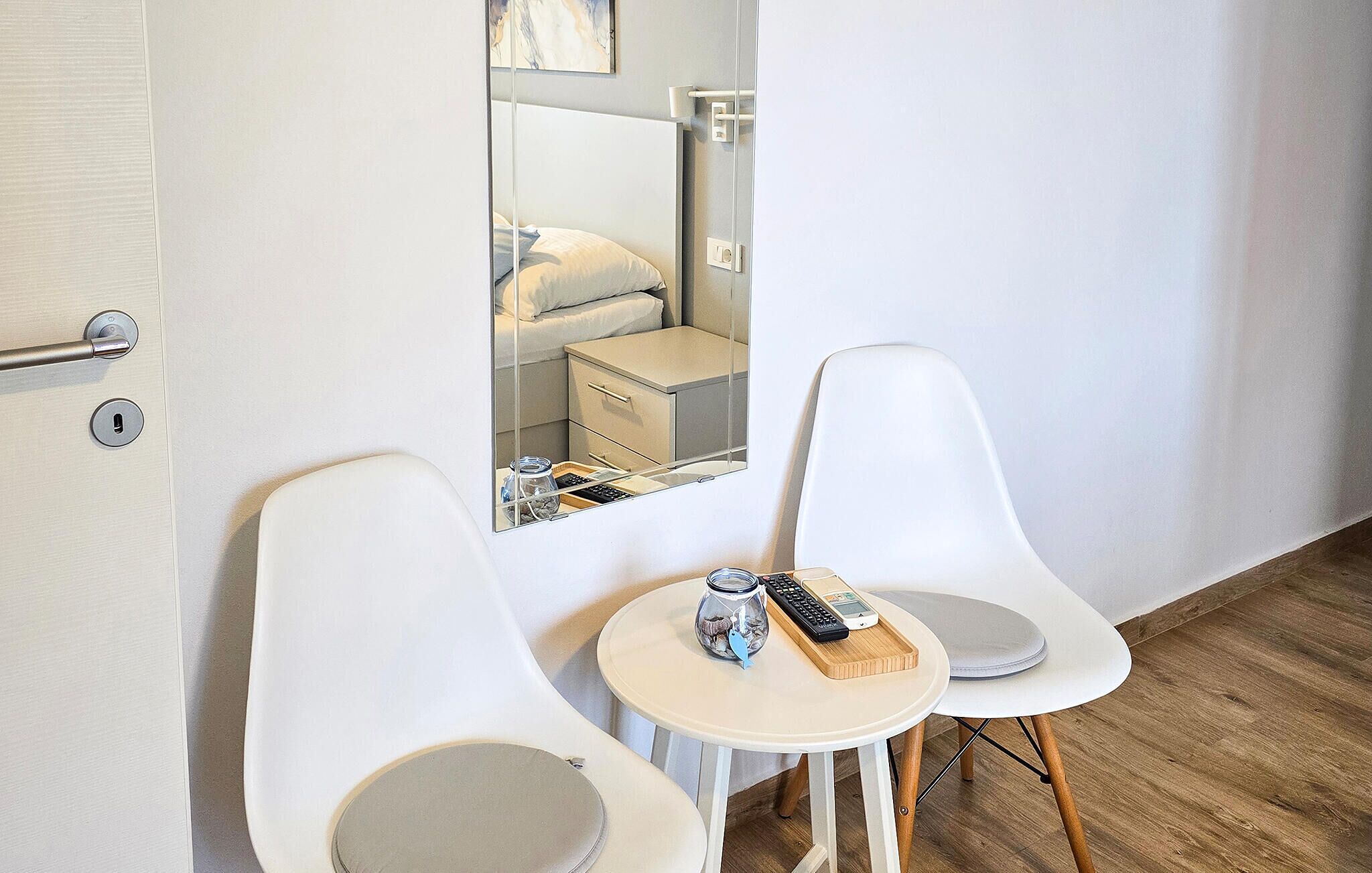 Een hotelkamer met een bed, een nachtkastje met een glazen pot en een afstandsbediening, twee witte stoelen en een klein rond tafeltje met een dienblad.