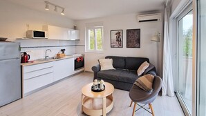 A modern kitchen with white cabinets, a microwave, and a red kettle. There's a grey sofa with cushions and a small round table with a tray. A window provides natural light and a view of greenery outside. - Nice apartment in Rab with WiFi (Rab)