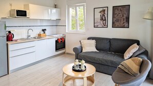 A modern kitchen with white cabinets, a microwave, and a stove. There is a grey sofa with cushions and a small round wooden table. A window provides natural light, and artwork is displayed on the wall. - Nice apartment in Rab with WiFi (Rab)