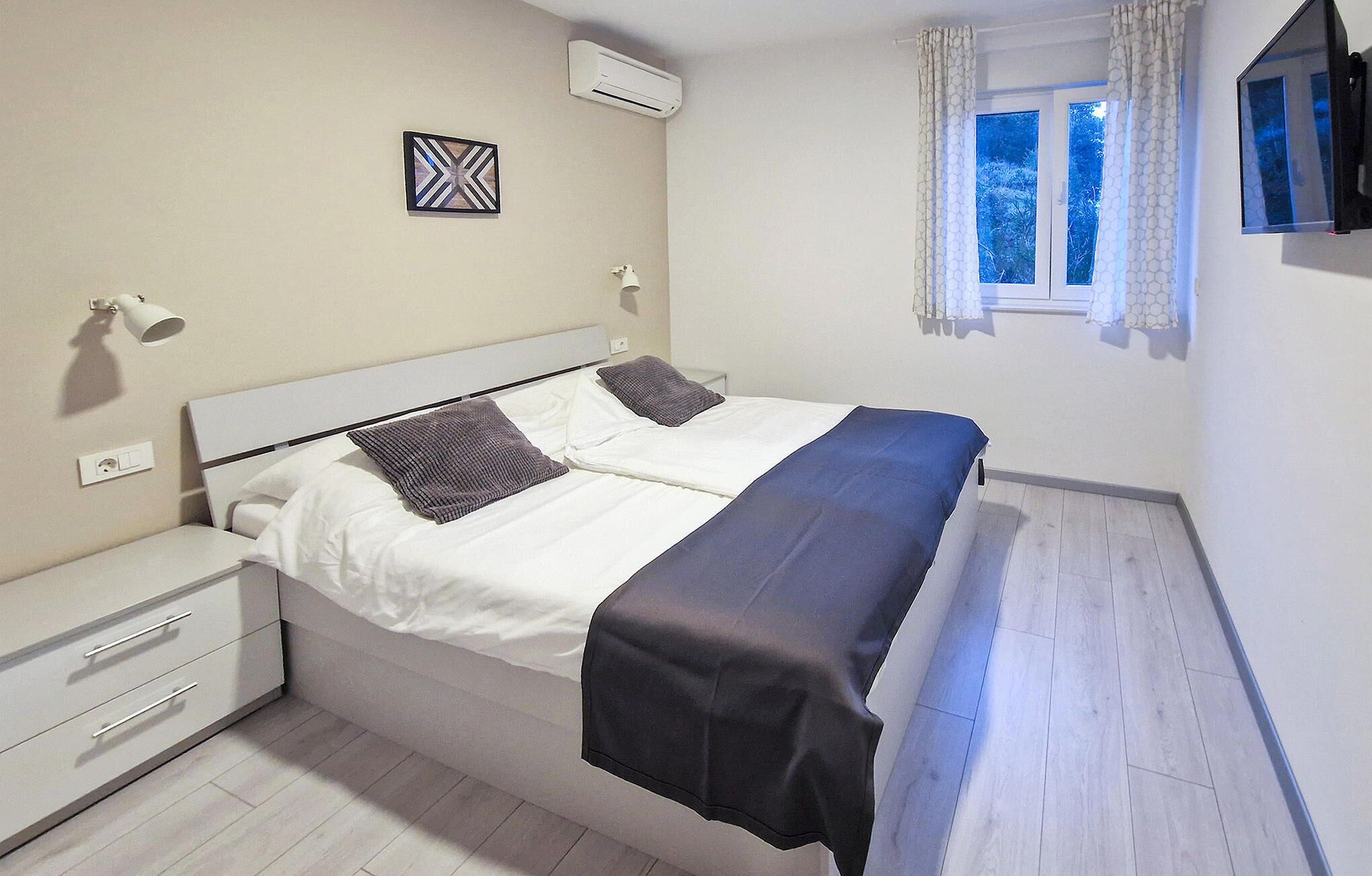 A bedroom with a bed, bedside tables, a wall-mounted air conditioner, a window with curtains, and a wall-mounted TV.