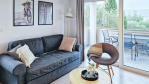 A modern living room with a dark grey sofa, a round wooden coffee table, and a unique chair with a curved backrest. There are two framed pictures on the wall, a floor lamp, and a view of the outdoors through a large window. - Nice apartment in Rab with WiFi (Rab)