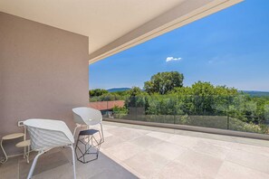 Un balcon avec deux chaises blanches et une petite table, d’où l’on peut admirer un paysage pittoresque, avec des arbres et une montagne lointaine.