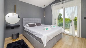 A modern bedroom with a grey upholstered bed, a round mirror, a bedside table with a charging station, and a view of greenery through the window. - Villa LA VIE with private pool (Garica)
