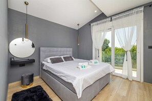 A modern bedroom with a grey upholstered bed, a round mirror, a bedside table, and a view of greenery through large windows. - Villa LA VIE with private pool (Garica)