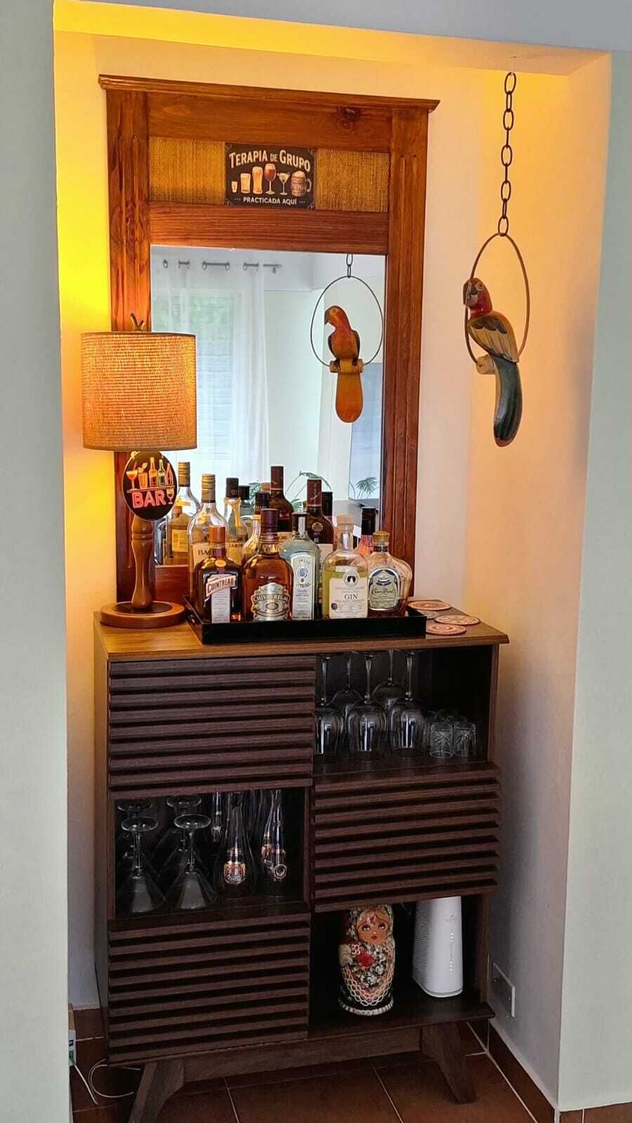 Una zona de bar con mostrador de madera, botellas, vasos y un adorno de pájaro colgante.