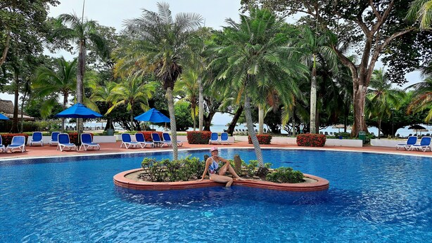 Zona de piscina con sillones reclinables azules y sombrillas, rodeada de exuberante vegetación y palmeras.