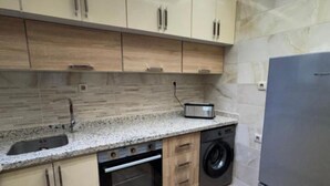 Een kleine keuken met houten kastjes, een inbouwoven, een wasmachine en een roestvrijstalen koelkast.