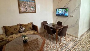 Een kamer met een bank, een eettafel met stoelen, een aan de muur bevestigde televisie en vloertegels met een patroon.