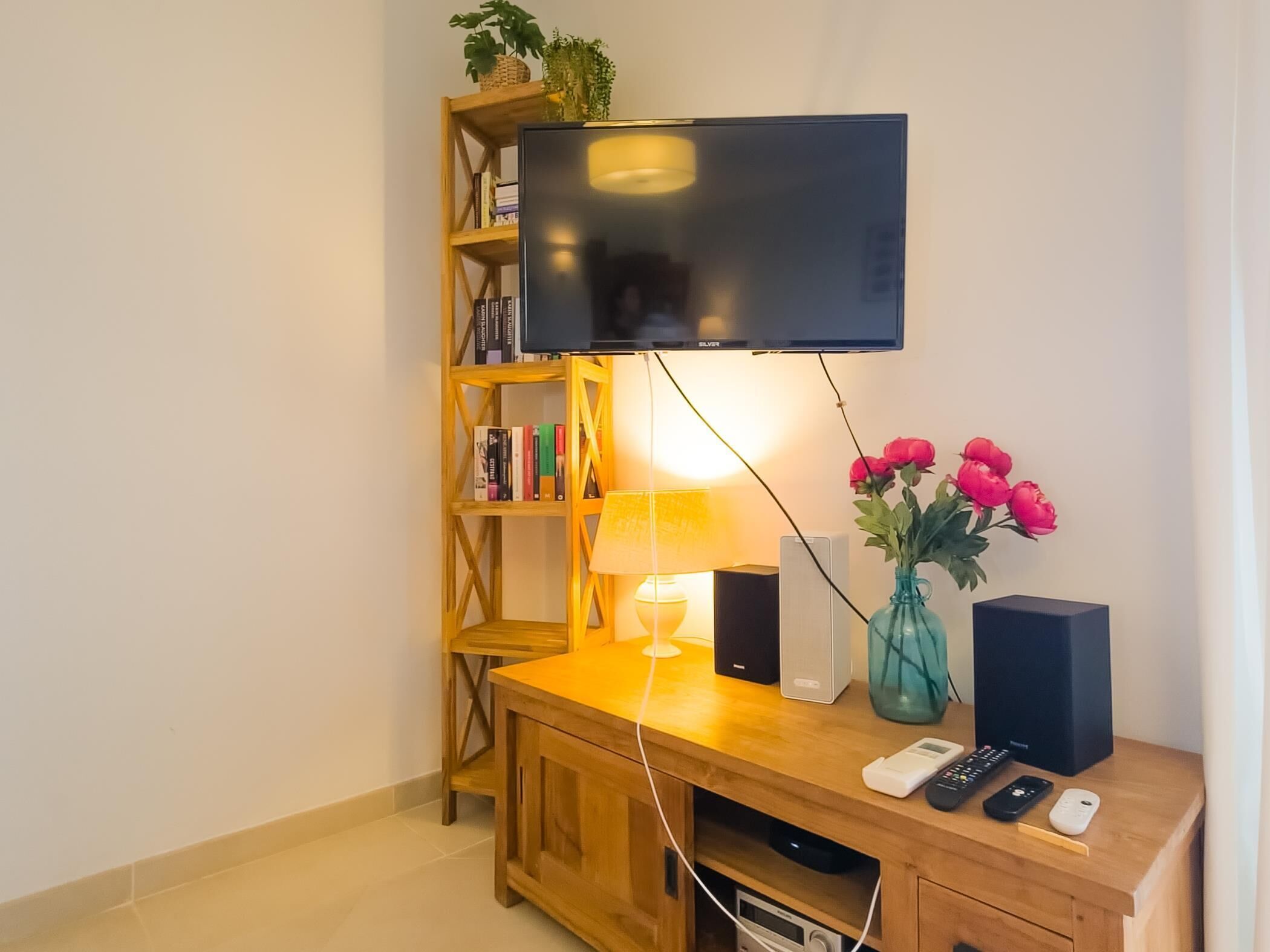 A wooden entertainment unit with a flat-screen TV, a lamp, a vase with flowers, and a speaker.