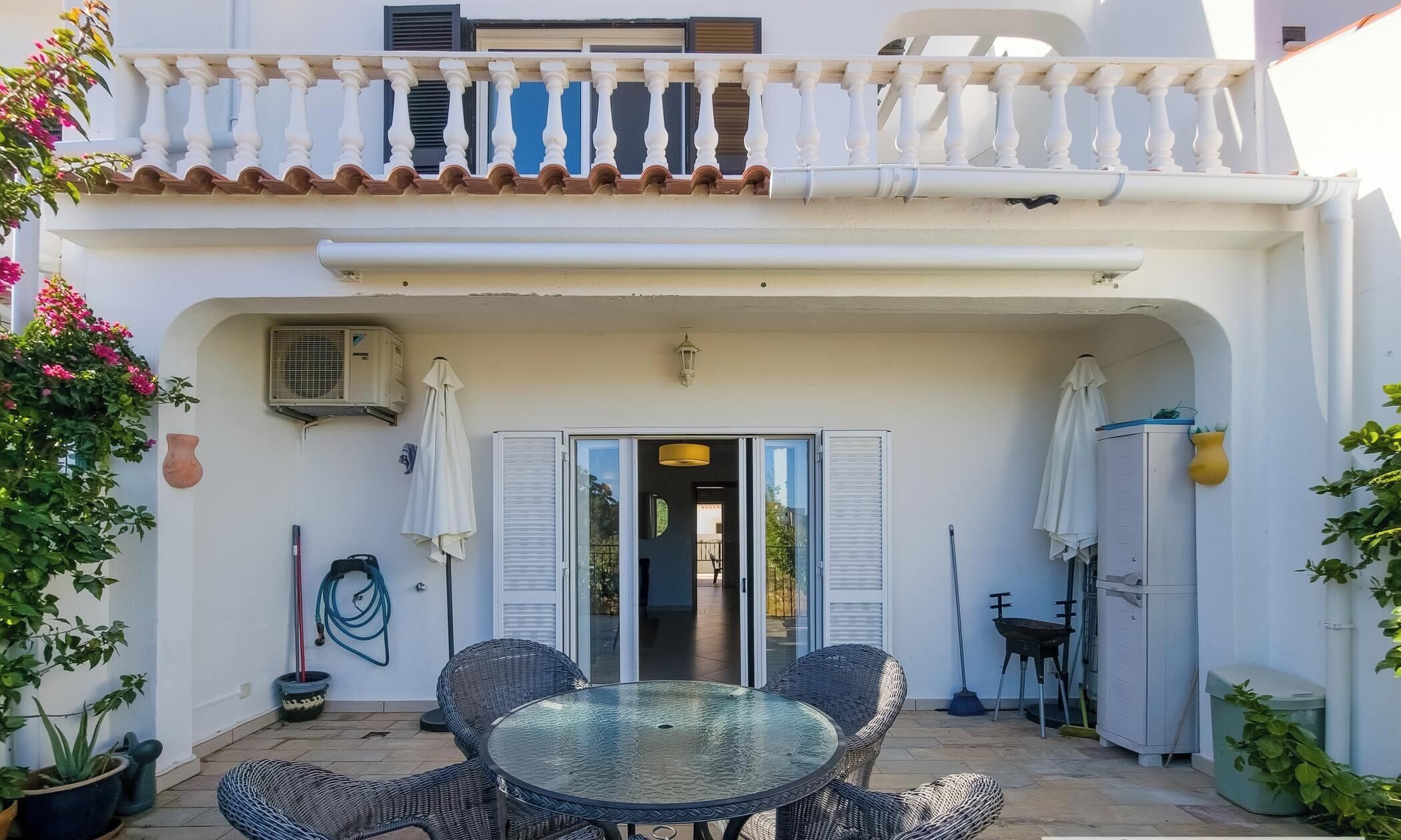 Une terrasse couverte avec une table en verre et des chaises en osier, un appareil de climatisation et des plantes en pot.