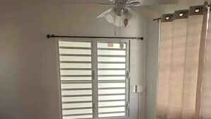 Une chambre à coucher comprenant un lit, un ventilateur de plafond, une fenêtre avec des stores, une table de chevet avec une lampe et un radiateur.