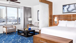 Une chambre d’hôtel avec un grand lit, une tête de lit en bois, deux tables de chevet avec des lampes, une chaise, une petite table avec un bol et un objet bleu, et une grande fenêtre donnant sur des immeubles.