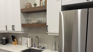 A modern kitchen with white cabinetry, a stainless steel refrigerator, and a wooden shelf above the sink. - Brand New! Downtown Stratford: The Blowes Inn 3C: 2 Bedrooms (Stratford)