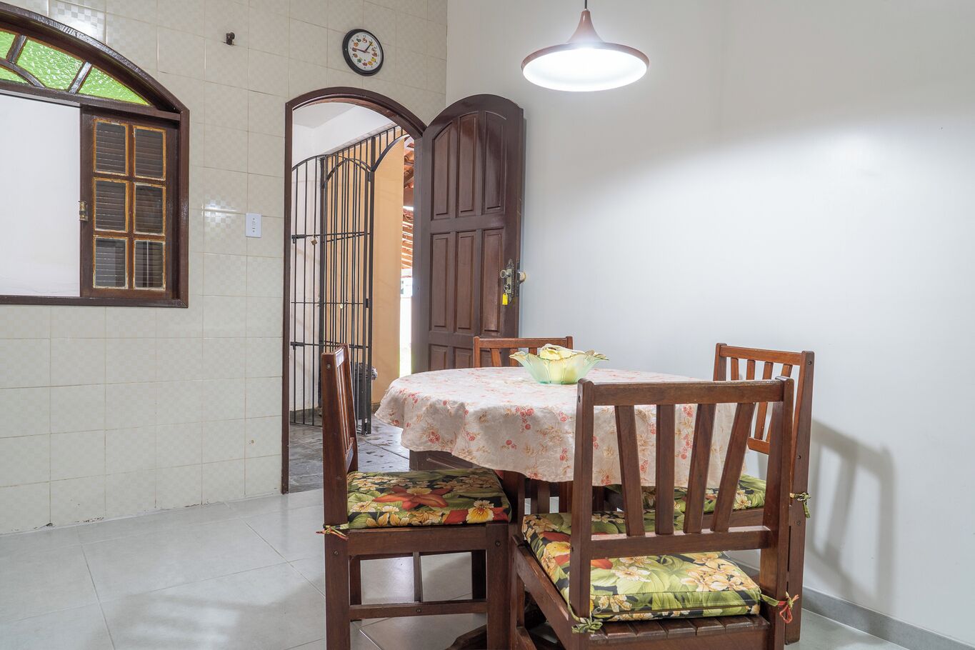 Un coin repas avec une table dressée pour quatre personnes, entourée de chaises en bois ornées de coussins à motifs floraux. Une horloge et un luminaire sont visibles sur les murs.
