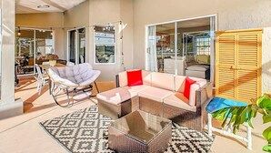 Une terrasse ensoleillée, meublée de meubles en osier, avec une table à plateau de verre, et d’où l’on peut admirer l’intérieur à travers des portes vitrées.