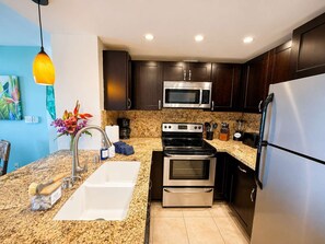 A modern kitchen with dark wood cabinets, stainless steel appliances, a granite countertop, and a hanging pendant light. - 2 Kings | Top Floor Oceanfront | Big Lanai | Views from the Top (Lahaina)