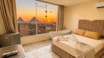 A hotel room with a bed, a small table with a remote control, a bathtub, and a view of pyramids at sunset.