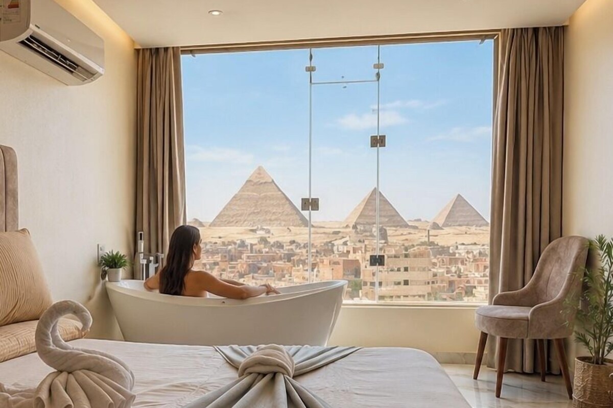 A hotel room with a bed, a bathtub, and a view of the pyramids.