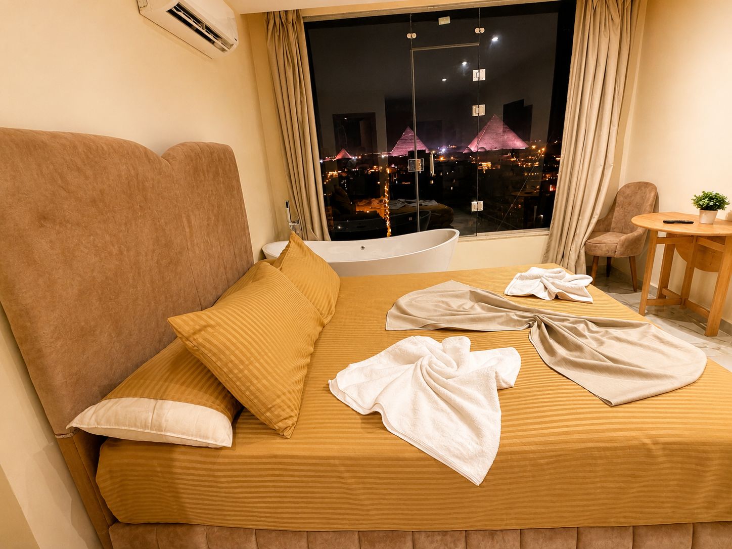 A hotel room with a bed, a chair, a small table, and a view of the pyramids at night.