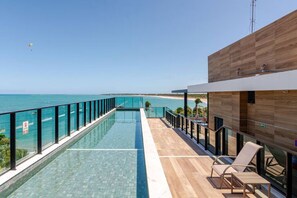 Une piscine à débordement moderne offrant une vue dégagée sur l’océan, avec une terrasse en bois et des chaises longues.