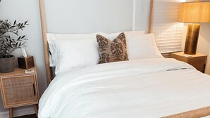 Ein ordentlich bezogenes Bett mit weißen Leinen, ein Holz-Kopfbrett und ein Nachttisch mit Lampe und Pflanze.