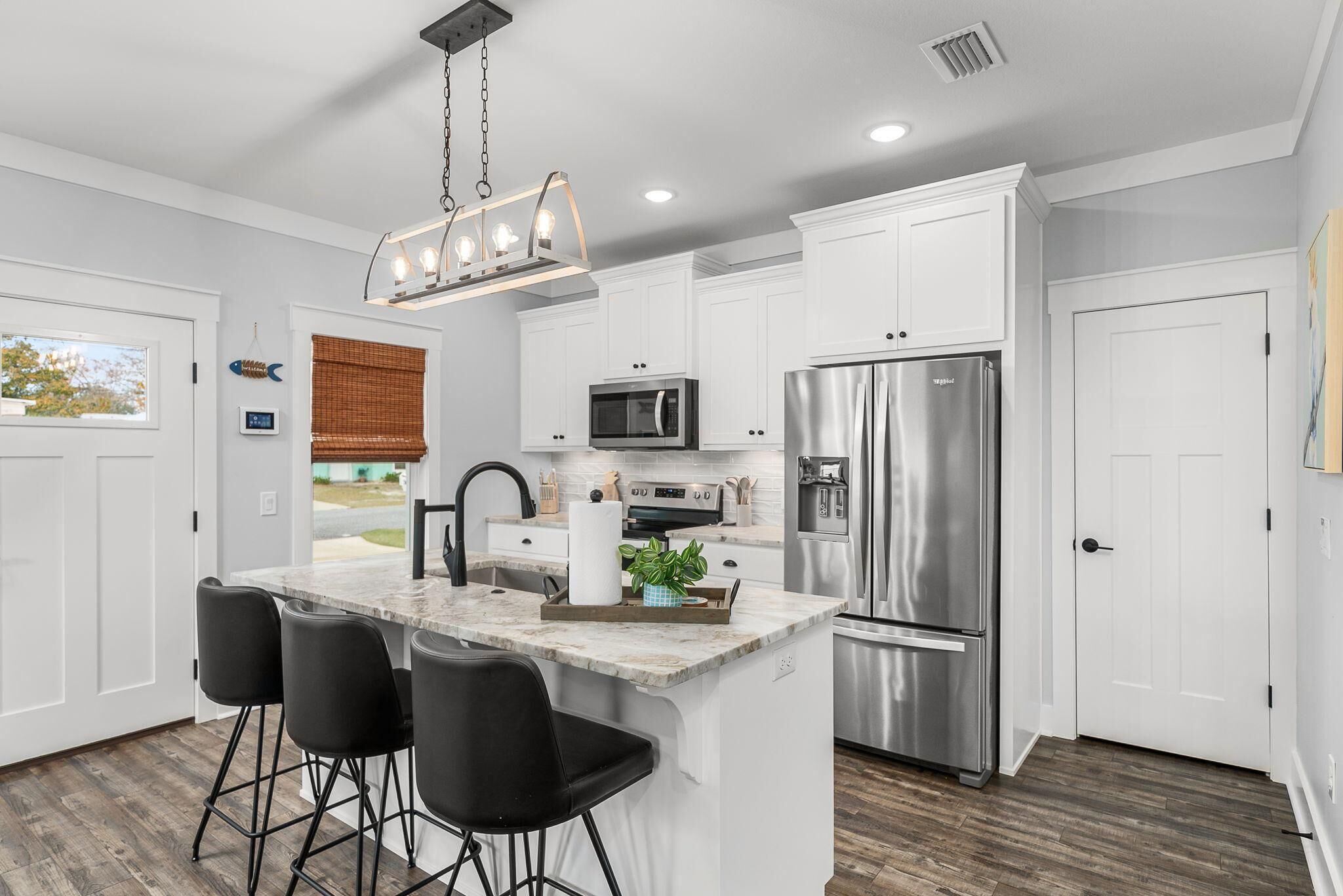 A modern kitchen with white cabinets, stainless steel appliances, a marble island with bar stools, and a hanging light fixture.
