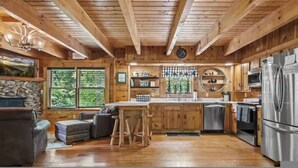 A rustic cabin interior with a wooden ceiling, stone fireplace, wooden bar, and modern appliances. - Black Bear Bungalow by Beyond Expectations (Gatlinburg)