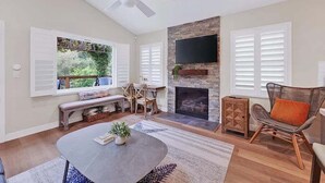 A spacious living room with a fireplace, a flat-screen TV mounted on a stone wall, and a ceiling fan. - Coastal Retreat in the Heart of Capitola Village - Steps from the Beach! (Capitola)