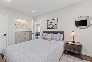A bedroom with a bed, a nightstand, a dresser, a mirror, and artwork on the walls. - Coastal Retreat in the Heart of Capitola Village - Steps from the Beach! (Capitola)