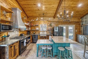 A spacious kitchen with wooden cabinetry, a central island with bar stools, stainless steel appliances, and a large window providing a view of the outdoors. - Frio Hills Hideaway | Creek | Mountain Views (Leakey)