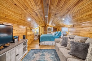 Ein gemütliches Blockhaus-Interieur mit Holzboden, einem Fernseher, einem Bett mit blauer Bettwäsche und einem Sofa mit gemusterten Kissen.